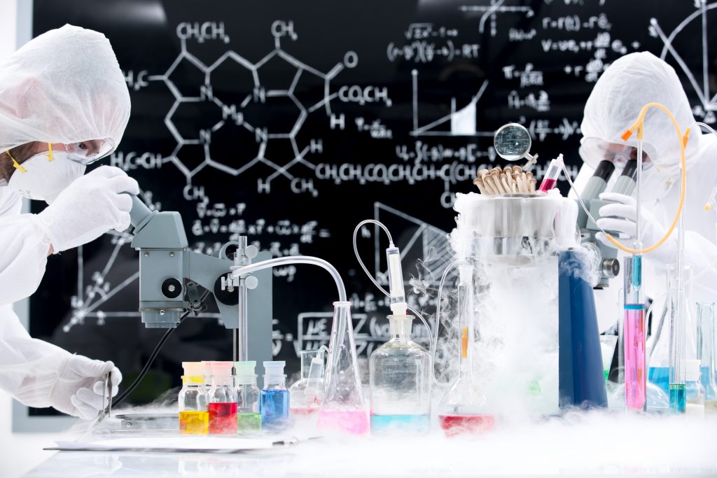 side-view of two people manipulating laboratory tools and colorful liquids on a lab table with a blackboard on the background