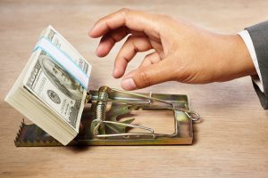 Image of a businessman reaching into a mousetrap for the bait, a stack of hundred-dollar bills