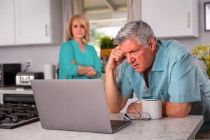 A concerned senior man looks at his laptop in the kitchen while his wife looks on from a distance.