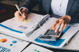 Image of a faceless businesswoman doing math on a calculator with an open binder of notes on her desk.