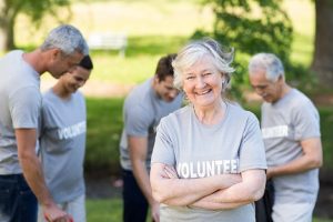 Image of a happy senior woman wearing a shirt that says “Volunteer” while others work in the background