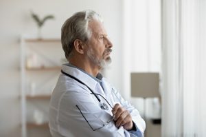 Image of an uncertain senior man staring out the window with his arms folded