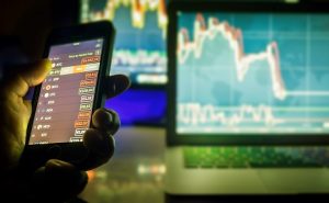 Image of a hand holding a phone used for day trading while a computer shows stock charts in the background