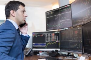 Image of a young man on the phone in front of multiple computer screens showing stock charts