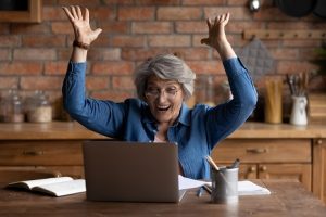 Image of a happy senior woman looking at her laptop with her hands in the air