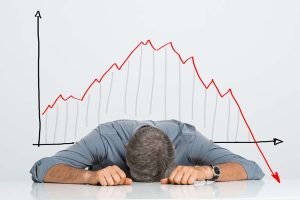 A frustrated investor laying his head on his desk in front of a falling stock chart