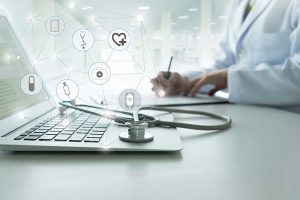 A doctor writing a note next to a stethoscope lying on a laptop