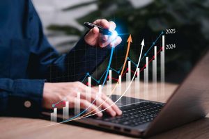 An investor sitting in front of a laptop with a pen in their hand. The pen is tracing an upward-facing chart.