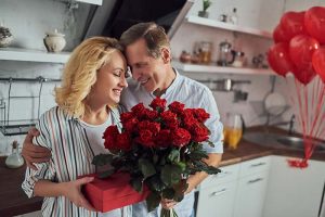 A middle-aged couple smiling and hugging while holding a box of chocolates and a bouquet of roses