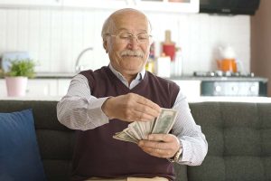 An elderly man sitting on a couch and smiling as he counts the dollar bills in his hands