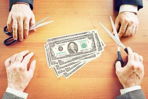 Two pairs of hands holding scissors around a pile of $100 bills on a table
