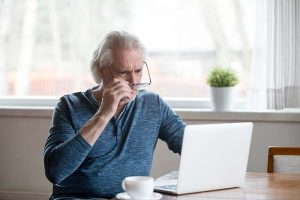 A retirement-age man removing his glasses as he looks as his laptop screen in disbelief