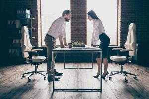 A man and a woman both dressed in business attire, standing at a desk and facing each other defiantly