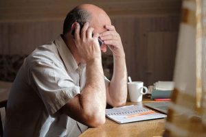 A distressed retirement-age man talking on the phone while sitting at a desk