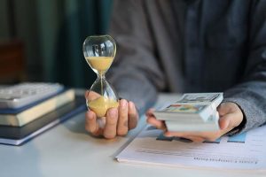 A businessman sitting at a desk with a stack of cash in one hand and an hourglass in the other